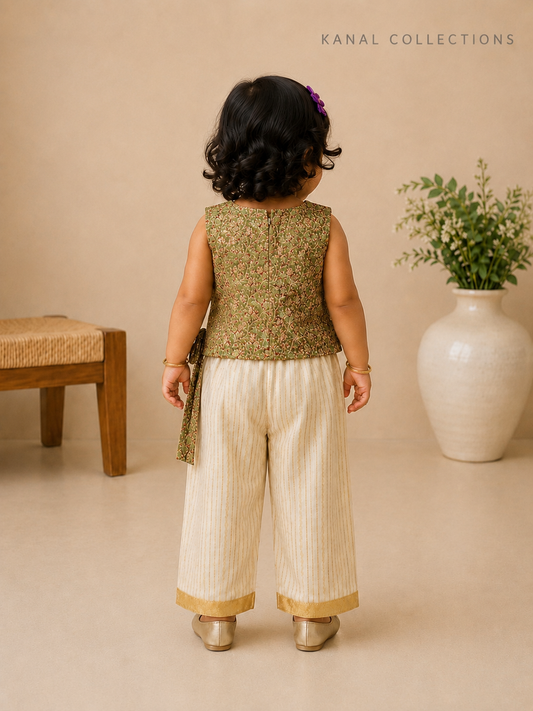 A toddler girl is shown from behind wearing a sleeveless olive green embroidered top and cream-colored pleated pants with gold trim, standing in a studio setting with a beige background, a wooden bench, and a large white vase with greenery.
