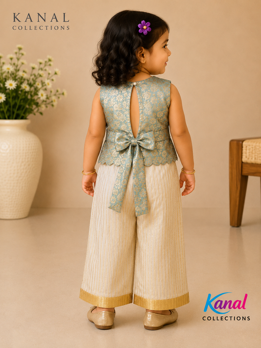 A toddler girl is shown from the back wearing a light blue-green and beige traditional outfit with a bow at the waist, standing in a warm beige room with a flower vase and wooden chair in the background. The brand logos 'KANAL COLLECTIONS' are visible in the corners.