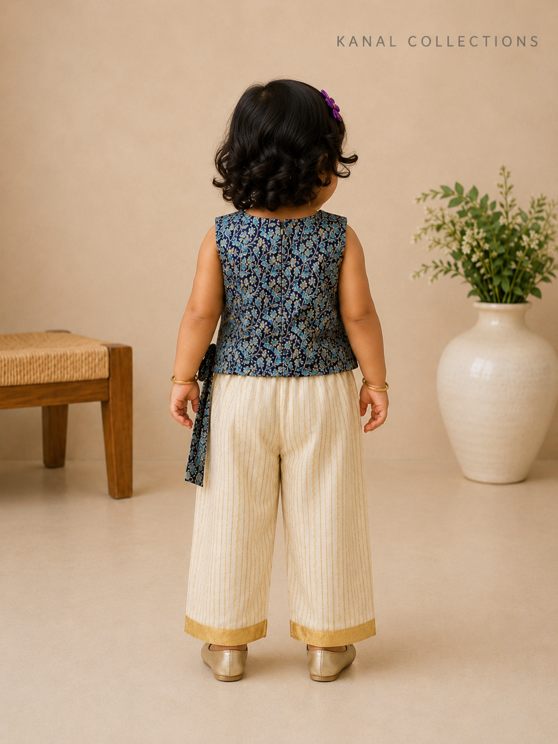 A toddler girl is shown from behind wearing a sleeveless navy blue patterned top and cream-colored pleated pants with gold trim, standing in a studio setting with a beige background, a wooden bench, and a white vase with greenery.