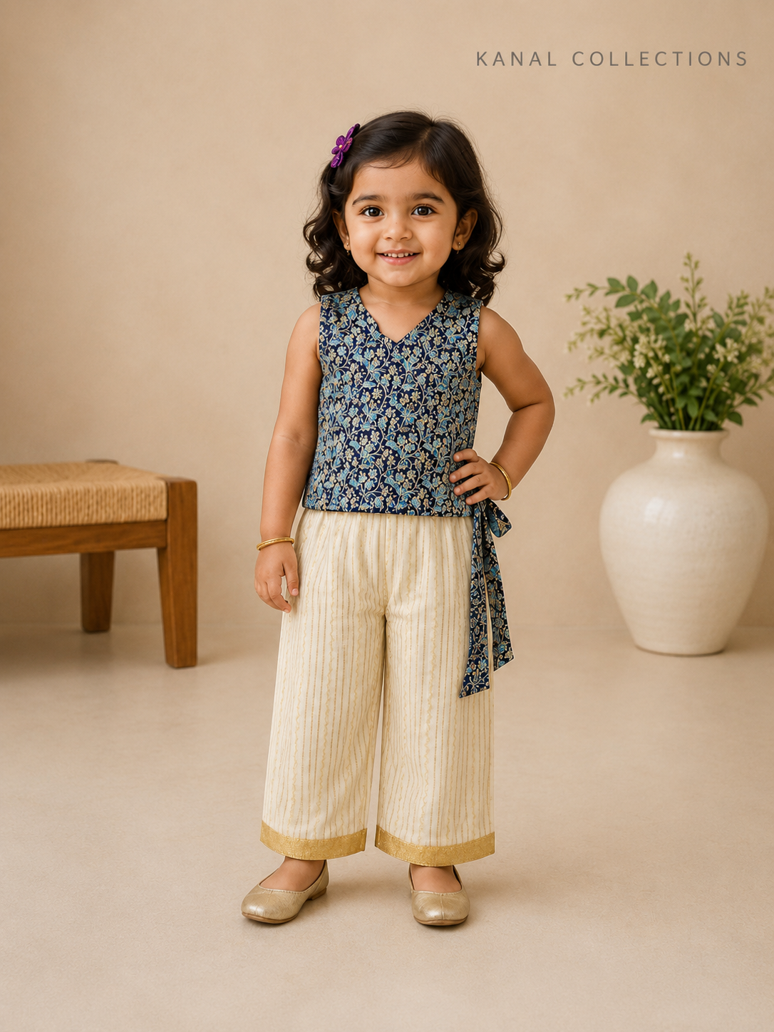 A young girl modeling a sleeveless navy blue floral printed top paired with cream-colored pleated wide-leg pants, standing in a studio setting with a beige background, a wooden bench, and a white vase with greenery. The brand name 'KANAL COLLECTIONS' is visible in the top right corner.