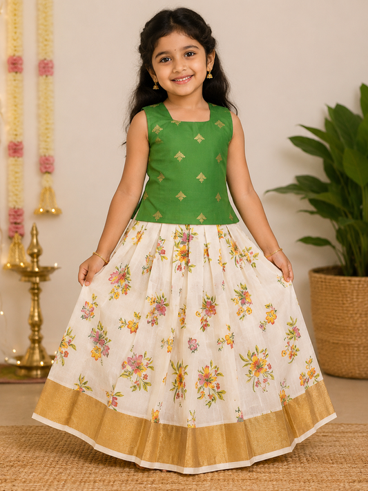 Little girl smiling in a traditional Indian ethnic outfit featuring a sleeveless green blouse with gold motifs and a cream floral pleated long skirt with a wide gold border.